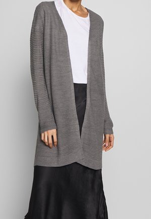 Kobieta w szarym prążkowanym kardiganie z odkrytym przodem, nałożonym na białą koszulkę, oraz czarną satynową spódnicę na tle jednolitego szarego tła.