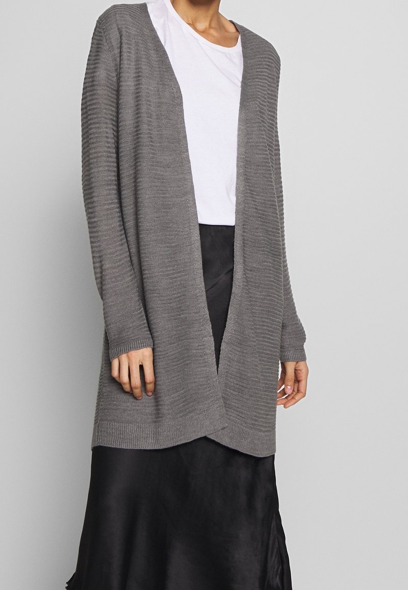 Femme portant un cardigan gris côtelé ouvert sur un t-shirt blanc et une jupe en satin noire devant un fond gris uni.