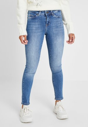 Frau trägt hellblaue Skinny-Jeans, weiße Sneakers und einen weißen gestrickten Pullover, steht vor einem einfarbigen Hintergrund.