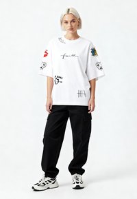 Balta per didelį t-shirt su įvairiais grafikos spaudiniais juoda ir spalvota, derinama su juodomis cargo kelnėmis ir baltais sportbačiais su juodais akcentais.