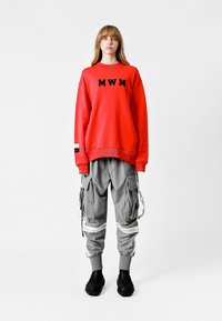 Sudadera roja con el bordado negro "MWM" en el pecho, combinada con pantalones cargo grises que tienen bolsillos y acentos blancos, y zapatos negros texturizados.