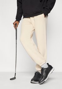 Μαύρα αθλητικά παπούτσια, μπεζ φόρμες και μαύρο φούτερ με κουκούλα. Το άτομο κρατάει ένα γκολφ κλαμπ, παρουσιάζοντας ένα αθλητικό και casual σύνολο.