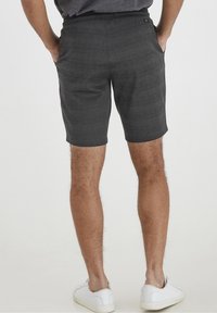 Pantaloni corti a quadri grigi con cintura elastica, tasche laterali e vestibilità sartoriale. Indossati con sneakers bianche su uno sfondo neutro.