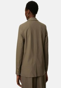 Osoba z krótkimi kręconymi włosami ubrana w oliwkowy żakiet w paski i pasujące spodnie, stojąca tyłem na tle jasnej, jednolitej ściany.