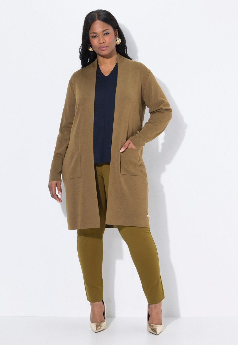 Frau trägt goldene Creolen, einen langen braunen Cardigan, ein marineblaues Oberteil, olivgrüne Hosen und goldene spitze High Heels, steht vor einer weißen Wand.