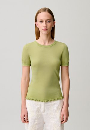 Joven mujer con cabello liso castaño claro lleva una camiseta ajustada de manga corta de color verde claro y pantalones blancos con estampado, de pie frente a un fondo liso.