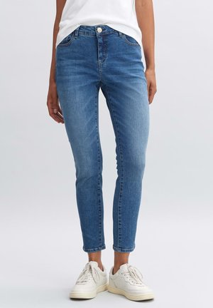 Persoon draagt nauwsluitende blauwe spijkerbroek en witte sneakers, staat tegen een effen lichte achtergrond met ontspannen handen langs het lichaam.