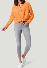 Chemise orange légèrement texturée à manches longues nouée à la taille, associée à un jean gris clair longueur cheville et des baskets blanches.