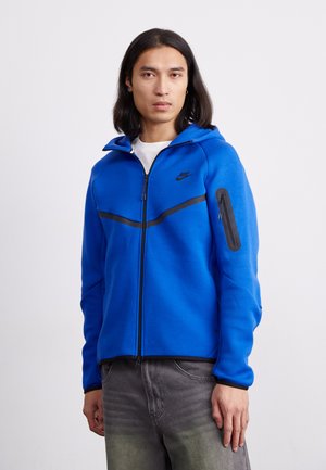 Blauwe ritsshirt met een zwarte diagonale streep over de borst; heeft een ritszak op de linkerarm en een verstelbare capuchon.