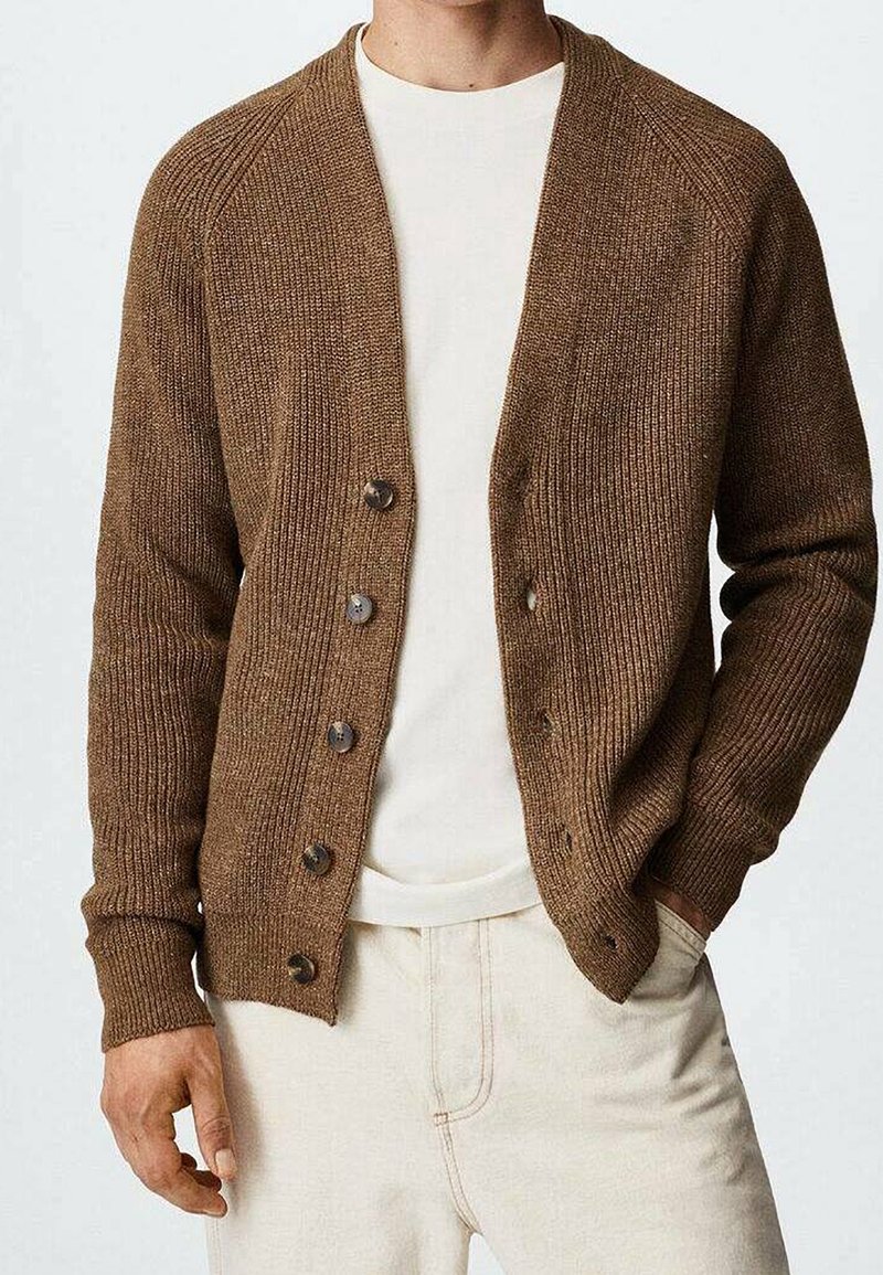 Gilet en tricot marron avec une fermeture à boutons, texture côtelée et manches longues. Porté sur un t-shirt de couleur claire et un pantalon crème.
