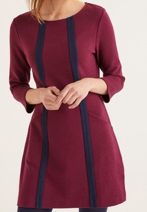 Žena nosící vínově červené šaty s dlouhým rukávem a dvěma vertikálními tmavě modrými pruhy na přední straně, ruce jemně sepnuté v pase.