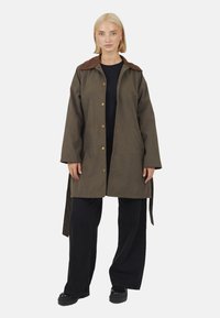 Cappotto marrone e verde oliva con chiusura a bottoni, colletto marrone e maniche lunghe. Presenta un taglio dritto e lacci laterali per la regolazione.