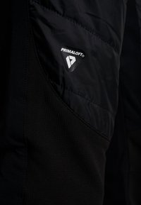 Primer plano de la tela de una chaqueta negra aislante con un parche del logo de Primaloft cosido en la sección acolchada.