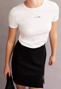 Osoba v bílém tričku s nápisem "Trvalá změna" a černé sukni s bočním rozparkem, která v jedné ruce drží černý předmět.