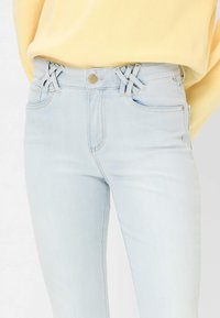 Jeans azzurri chiaro a vita alta con passanti doppi incrociati unici, indossati con un maglione giallo infilato in vita.