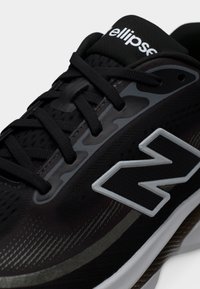 Primo piano di una scarpa da corsa New Balance nera con logo "N" grigio e bianco e lacci neri su sfondo bianco semplice.