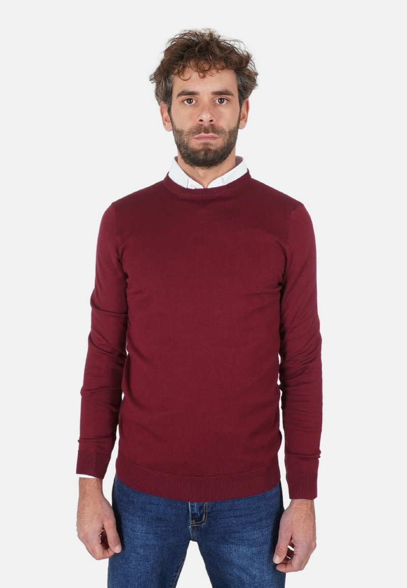 Maglione bordeaux lavorato a maglia con scollo rotondo, polsini e orlo a coste. Indossato sopra una camicia bianca con colletto, abbinato a jeans blu.