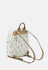 Liten vit ryggsäck med blommönster, beige justerbara remmar, ett handtag upptill och en zip-ficka på baksidan.
