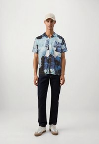 Trumpomis rankovėmis marškiniai mėlynos ir juodos abstraktaus rašto; audinys atrodo lygus. Derinama su tamsiomis džinsinėmis kelnėmis ir baltomis sportinėmis avalynėmis; dėvima smėlio spalvos kepurė.