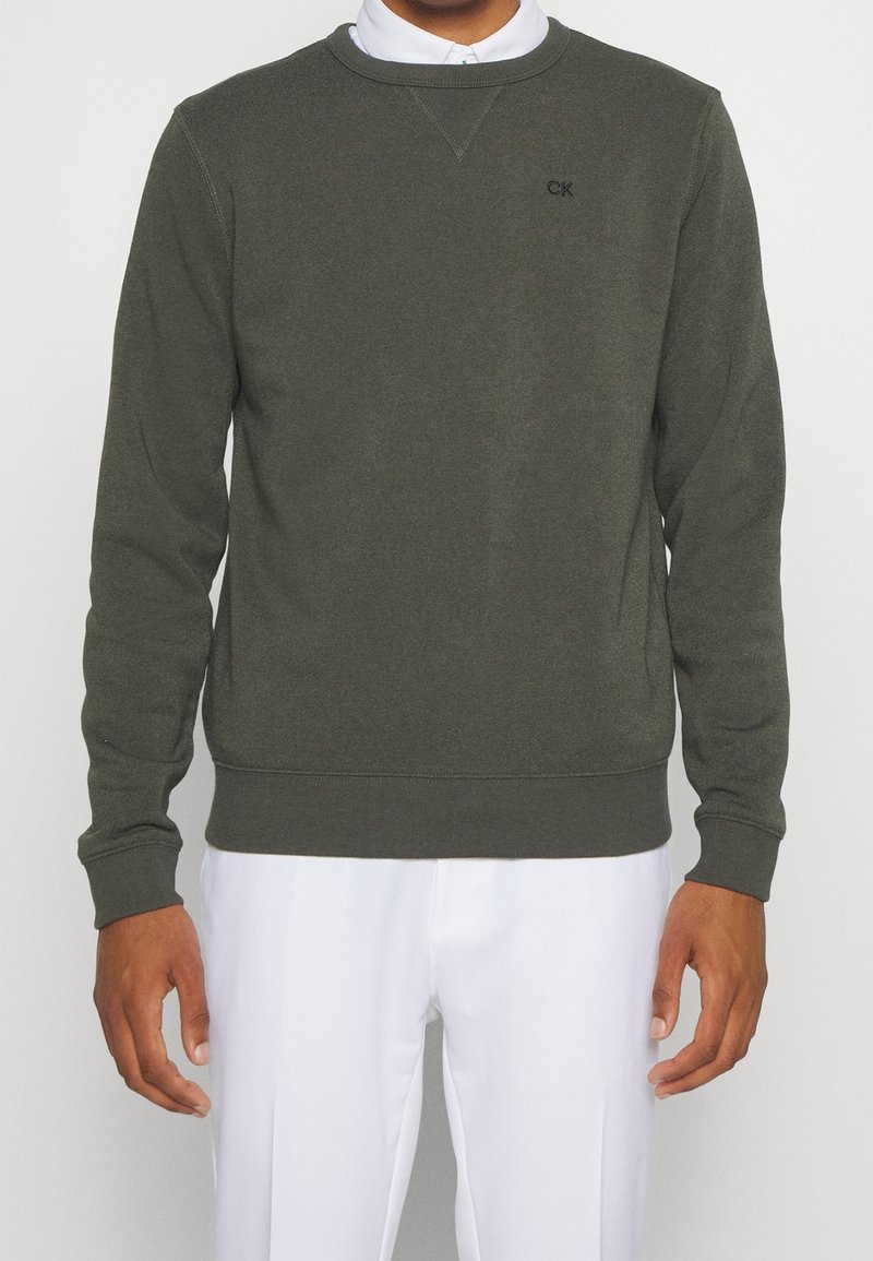 Homme portant un pull-over à manches longues vert foncé avec petit logo "CK" et pantalon blanc, debout devant un fond clair uni.
