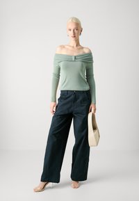 Off-shoulder ljusgrön topp kombinerad med mörkblå vida byxor. Modellen håller en beige handväska. Minimalistisk design med mjuka texturer.