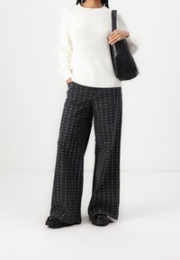 Pull en maille blanche associé à un jean noir à jambes larges orné de pois blancs. Le mannequin tient un sac à épaule noir et porte des chaussures noires épaisses.