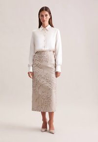 Gonna beige ricamata con motivo floreale, abbinata a una camicetta bianca con bottoni. L'outfit presenta una silhouette aderente e una texture liscia.