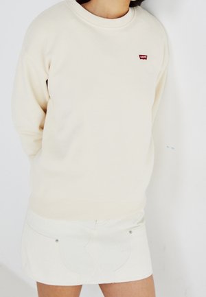 Personne portant un sweat-shirt Levi's crème et une jupe en denim blanche, debout les mains derrière le dos devant un fond clair uni.