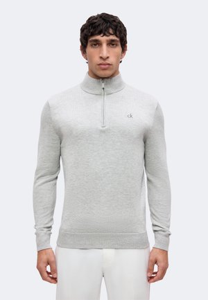 Giovane uomo che indossa un maglione grigio chiaro con mezza zip e piccolo logo "ck" sul petto, e pantaloni bianchi su sfondo semplice.