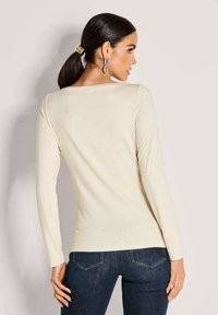 Langärmliges Creme-Oberteil mit geripptem Textur, weiter Ausschnitt, kombiniert mit dunkelblauen Jeans. Modell von hinten mit einem Pferdeschwanz.