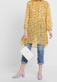Tunica gialla a fantasia floreale con maniche lunghe e scollatura arricciata, abbinata a jeans blu arrotolati e scarpe con tacco a blocco in suede viola.