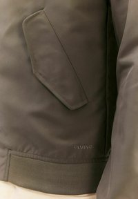 Olive green bomber jacket with a textured finish, featuring a diagonal pocket, ribbed hem, and embroidered brand logo at the bottom.