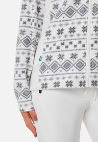 Polarowa kurtka w kolorze białym z szarymi geometrycznymi wzorami i wzorami płatków śniegu. Posiada zamek błyskawiczny z przodu, boczne kieszenie oraz małą niebiesko-żółtą metkę.