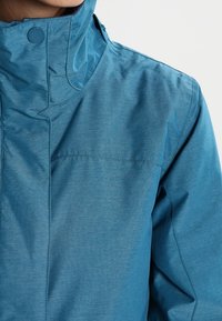 Tealblaue Jacke mit hohem Kragen, dezenter Textur, Druckknopfverschluss und gestickten Details, geeignet für den Outdoor-Einsatz.