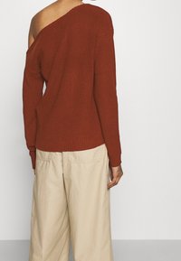 Knitted rust-colored sweater with an off-shoulder design, ribbed texture, and long sleeves, paired with light beige wide-leg pants.