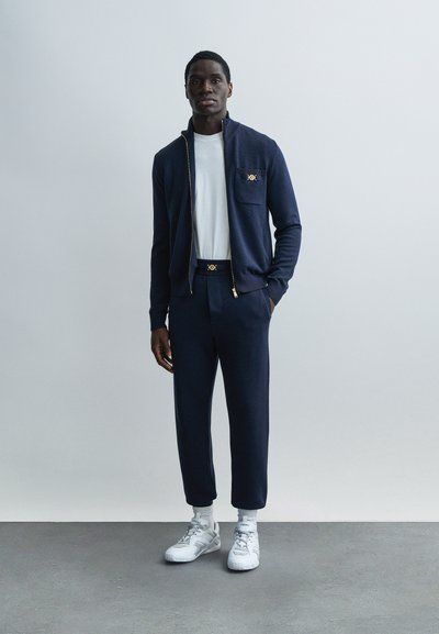 Marineblauw vest met rits en gouden accenten en zak, gecombineerd met bijpassende taps toelopende broek en witte sneakers. Gladde stofstructuur overal.