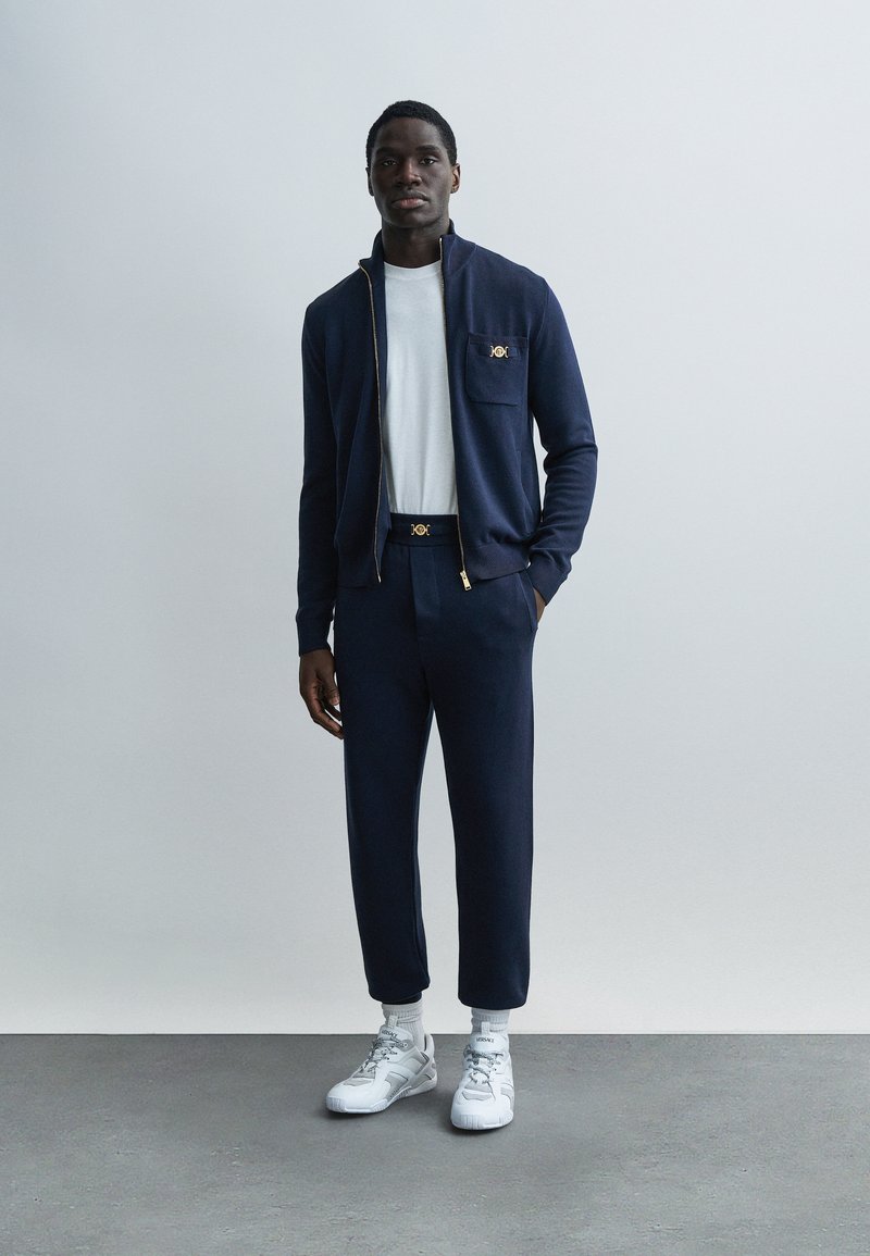 Marinblå zip-up jacka med gulddetaljer och ficka, kombinerad med matchande avsmalnande byxor och vita sneakers. Mjuk tygstruktur överallt.