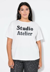 Biała bawełniana koszulka z krótkimi rękawami z odważnym czarnym napisem "Studio Atelier". Noszona z czarnymi spodniami w wzór z cekinów.