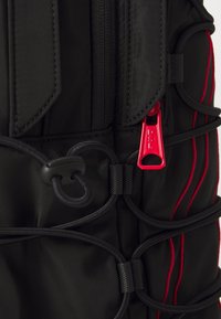 Sac à dos noir avec des bandes d'accent rouge, doté d'un tirant de fermeture éclair rouge bien visible, de détails en cordon élastique et d'une texture de tissu durable.