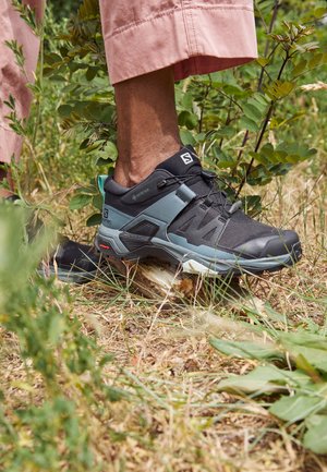 Chaussures de randonnée noires et bleu sarcelle avec un design robuste en mesh et matériaux synthétiques, posées sur une bûche au milieu de l'herbe et du feuillage.