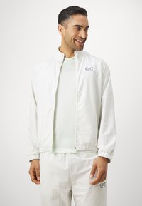 Jaqueta branca com fecho éclair e um padrão de grelha subtil, apresentando punhos elásticos e um logo no peito, combinada com uma blusa verde-clara.