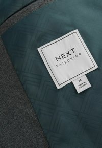 Maßgeschneiderter Jackenstoff mit grauem Wolluntergrund und grünem Musterteil. Verfügt über ein weißes Etikett mit der Aufschrift "NEXT Tailoring", Größe M.
