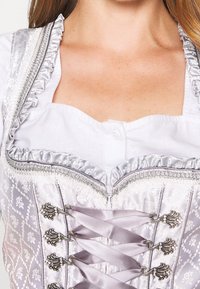 Frau in einer weißen Bluse und einem silbernen Seidenkorsett mit floralem Stickerei, Rüschensaum und dekorativen Metallverschlüssen mit Bändern.