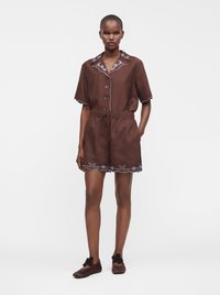 Femme portant un haut marron à manches courtes avec des boutons et un short assorti avec une broderie bleue, associé à des ballerines marron avec des nœuds noirs.