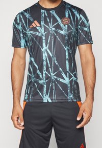 Musta urheilupaita, jossa on sininen abstrakti geometrinen kuvio, pyöreä kaulus, lyhyet hihat ja FC Bayern Münchenin logo rinnassa.