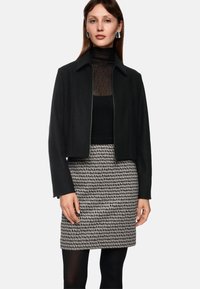 Schwarze kurzgeschnittene Jacke mit glatter Textur, kombiniert mit einem transparenten schwarzen Oberteil mit Polka-Dots und einem gemusterten schwarz-weißen Rock.