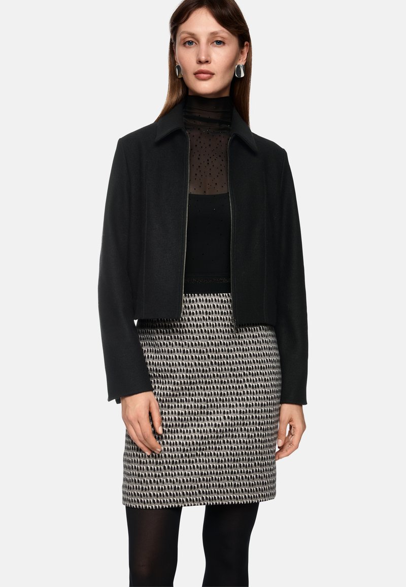 Schwarze kurzgeschnittene Jacke mit glatter Textur, kombiniert mit einem transparenten schwarzen Oberteil mit Polka-Dots und einem gemusterten schwarz-weißen Rock.