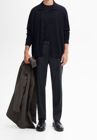 Navy cardigan with buttons, worn over a black shirt and dark trousers. Holding a brown overcoat, and wearing black shoes. Simple design, smooth texture.