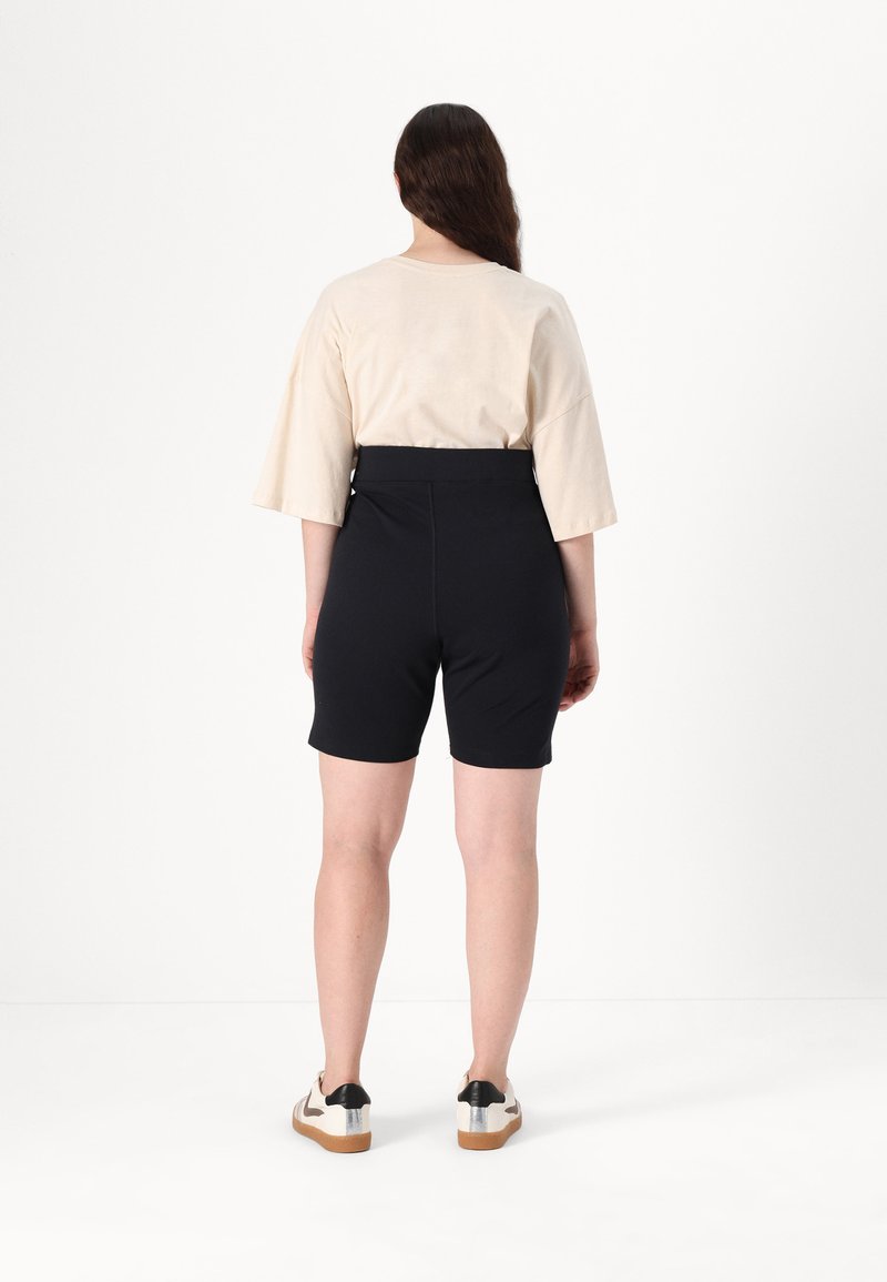 Zwarte op maat gemaakte shorts met een hoge tailleband, gecombineerd met een beige top met wijde mouwen. De schoenen zijn wit met zwarte en zilveren accenten.