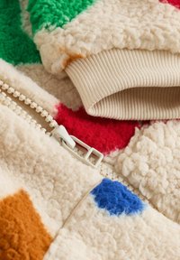 Fleece-takki, jossa on kermavärinen pohja ja värikkäitä läiskiä vihreänä, punaisena, ruskeana ja sinisenä. Mukana valkoinen vetoketju ja ribbikuitupäät.